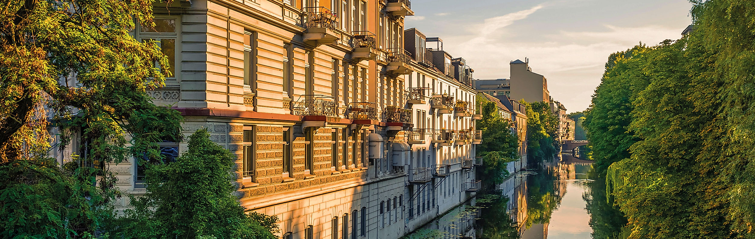 Jugendstilfassaden am Alsterkanal in Hamburg bei Sonnenuntergang, gesäumt von Bäumen und Wasserreflexionen